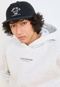 Sweatshirt gris avec capuche, arborant un logo imprimé noir "STUDIO EDITION" sur le devant. Casquette noire avec broderie blanche d'un canard et texte.