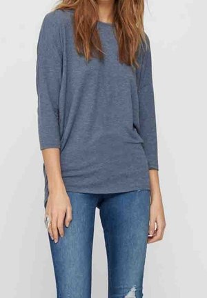 Femme portant un haut bleu ample à manches longues et un jean bleu déchiré ajusté, debout devant un fond clair uni.