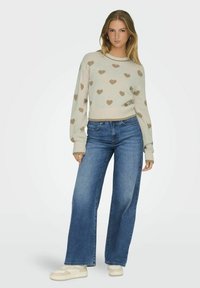 Beiger Pullover mit braunen Herzmustern, schmalen Bündchen und rundem Halsausschnitt, kombiniert mit hochtaillierten, blauen weiten Jeans und cremefarbenen Sneakers.