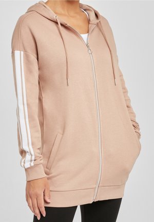 Sweat à capuche long beige avec fermeture éclair, bandes blanches sur les manches, poches avant et cordon de serrage à la capuche, porté sur un haut blanc et un pantalon noir.