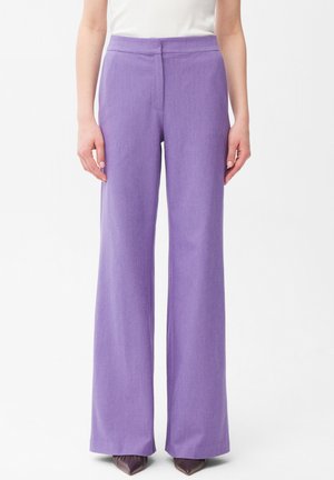 Proste, wysokowaistowe fioletowe spodnie noszone z brązowymi, spiczastymi butami i białą bluzką, pokazane na stojącej osobie na białym tle.