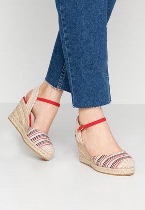 Füße, die Keil-Espadrilles mit roten Knöchelriemen und gestreiftem Stoff tragen, kombiniert mit blauen Cropped-Jeans vor einfachem Hintergrund.
