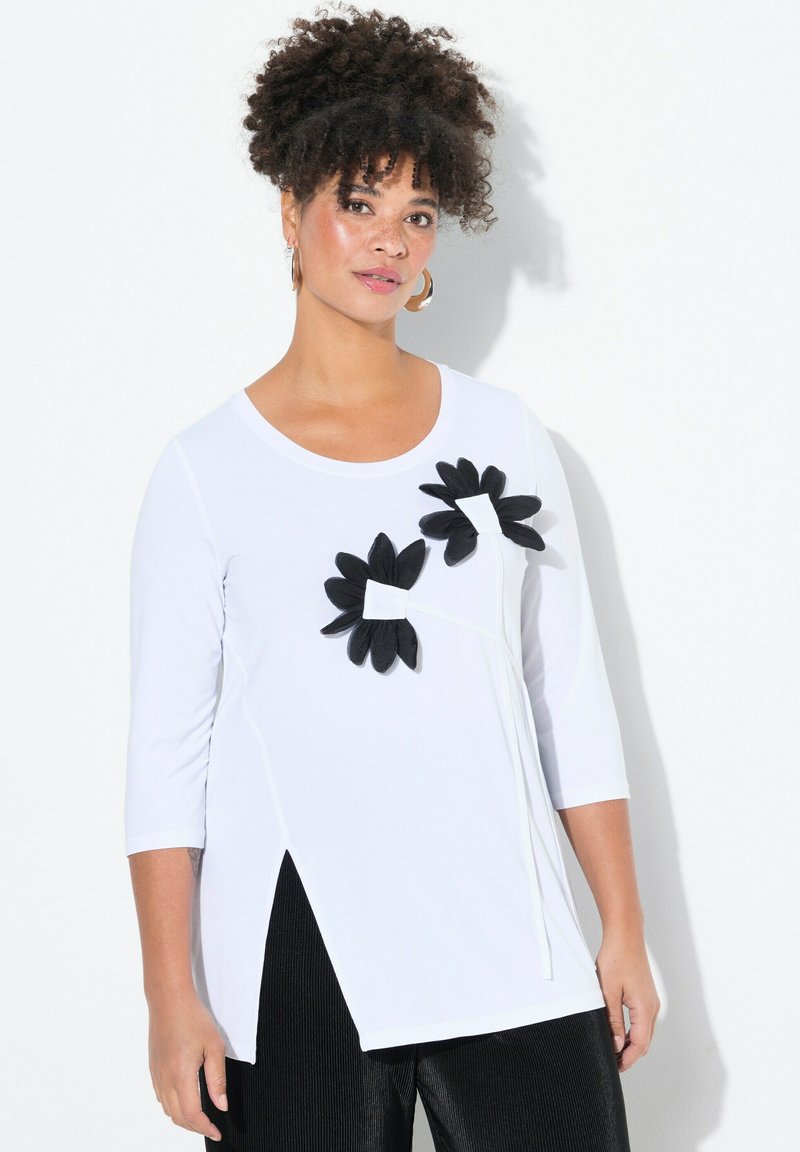Top bianco con maniche tre quarti, caratterizzato da due decorazioni floreali nere e uno spacco laterale. Materiale morbido con vestibilità comoda.