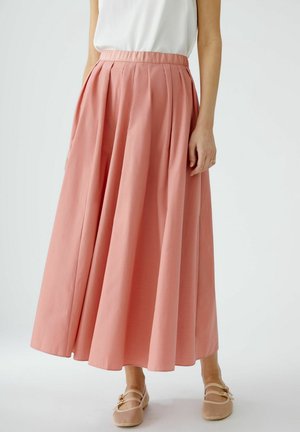 Vrouw draagt een hooggetailleerde, geplooide midi rok in koraalroze met beige platte schoenen op een effen achtergrond.