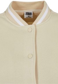 Béžová bomber bunda s ribbed límcem a bílými proužky. Zapínání na dva kulaté, světle béžové druky. Měkká, strukturovaná tkanina.