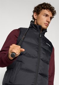 Μαύρο επενδυμένο γιλέκο με υψηλό γιακά, κλείσιμο με φερμουάρ και λεπτομέρεια λογοτύπου, φορεμένο πάνω από ένα μπορντό μπλουζάκι με μακριά μανίκια, κρατώντας ένα μπαστούνι γκολφ.