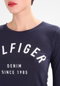 Námořnická modrá mikina s dlouhým rukávem vyrobená z bavlny, s velkým bílým textem sítotiskem "TOMMY HILFIGER DENIM SINCE 1985" napříč hrudníkem.