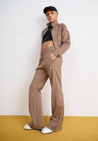 Brūns sporta treniņtērps ar ar rāvējslēdzēju aprīkotu jaku un biksēm ar elastīgu jostasvietu, apvienots ar melnu crop top un baltām sporta kurpēm uz dzeltena grīdas.