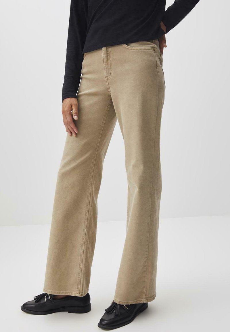 Beige weit geschnittene Hosen aus Baumwolle mit strukturierter Oberfläche, die über Nahtdetails und seitliche Taschen verfügen. Getragen mit schwarzen Slippern.