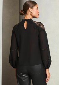Blusa nera con dettagli in pizzo sulle spalle, schiena a chiave e maniche lunghe a sbuffo. Abbinata a pantaloni in pelle nera aderenti. Texture liscia.