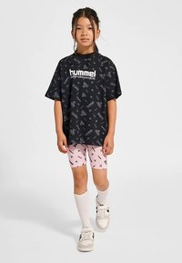 Sort oversized t-shirt med hvidt Hummel-logo og gentaget mønster, kombineret med lyserøde shorts med sorte detaljer og hvide sokker.