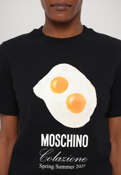 Zwarte T-shirt met een grote afbeelding van een gebakken ei met twee dooiers en de tekst "Moschino Colazione Lente Zomer 2025" eronder.