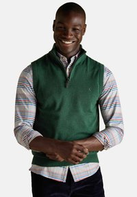 Šypsosi vyras, vilkintis žalią be rankovių pusiau užtrauktuką megztinį ant daugiaspalvio languoto marškinėlio su sukeltomis rankovėmis ir tamsiomis kelnėmis.