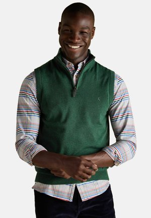 Lächelnder Mann in einem grünen ärmellosen Pullover mit halbem Reißverschluss über einem mehrfarbigen Karoshemd mit hochgerollten Ärmeln und dunklen Hosen.