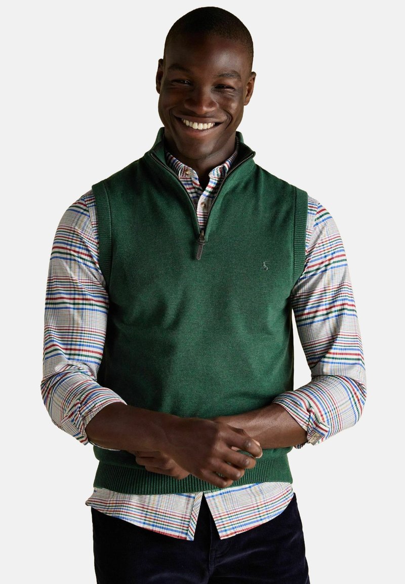 Šypsosi vyras, vilkintis žalią be rankovių pusiau užtrauktuką megztinį ant daugiaspalvio languoto marškinėlio su sukeltomis rankovėmis ir tamsiomis kelnėmis.