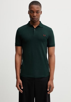 Homme portant un polo à manches courtes vert foncé avec un petit logo laurier rouge sur la poitrine gauche, debout devant un fond uni.