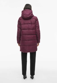 Bordo puffer jakna s kapuljačom, s pogodnim dizajnom, do koljena i od mekog, glatkog materijala. Vidljive crne hlače i štikle.