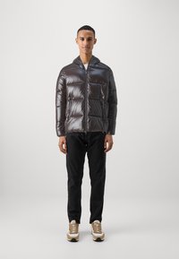 Donkergroene pufferjas met een glanzende afwerking, volledige rits, capuchon, twee zijzakken en horizontaal gestikte vulling. Gemaakt in combinatie met zwarte broek.