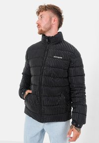 Zwarte pufferjack met een gestikte textuur, hoge kraag, ritssluiting en zijzakken. Gemaakt van glanzend materiaal met detail van het merklogo.