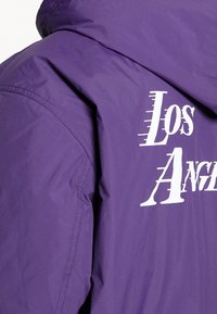 Lila jacka med huood, med stor vit text "Los Angeles" på baksidan. Slät tyg med matt finish och diskreta sömmar.