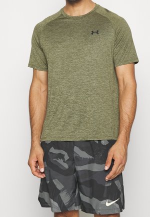 Mand iført olivengrøn Under Armour kortærmet skjorte og sort-grå camouflage Nike-shorts, stående med armene afslappet langs siden.