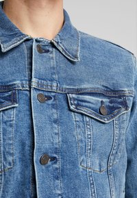 Ljusblå denimjacka med spetsig krage, två bröstfickor med knappstängning och synliga söm detaljer.