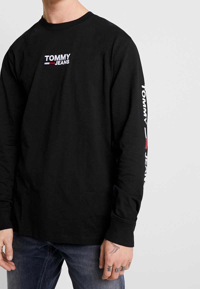 Svart långärmad T-shirt med "TOMMY JEANS"-logotyp i vitt och rött, med text längs vänster ärm. Tillverkad av bomull, med rund halsringning.
