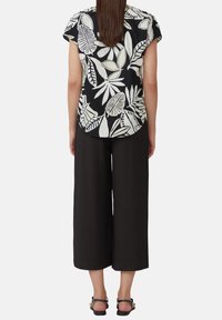 Svart blus med kort ärm och vita bladmönster, i kombination med vida svarta byxor. Materialet ser strukturerat ut; sandalerna har en enkel design.