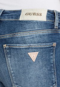 Vaqueros de denim con un lavando azul claro, que presentan una etiqueta texturizada de Guess en la cintura trasera y un parche con logo en forma de triángulo en el bolsillo trasero.
