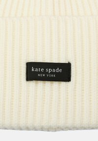 Kermavaahto ribbed-kudottu pipo, jossa musta kangastunnus, jossa lukee "kate spade NEW YORK" valkoisella tekstillä. Pehmeä rakenne, sylinterimäinen muoto.