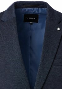 Donkerblauwe blazer met een gestructureerde buitenkant, satijnen voering, geknipte revers en een klein metallic accent op de kraag dat het merksymbool laat zien.