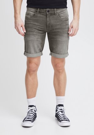 Homme portant un short en denim gris retroussé, des chaussettes blanches montantes et des baskets montantes noires, debout devant un fond uni.