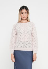 Jasnoróżowy sweter dzianinowy z wzorem w zygzak, z bufiastymi rękawami i prążkowanym wykończeniem. Noszony na ciemnoniebieskiej spódnicy. Gładka tekstura.