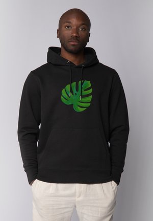 Mann trägt schwarzen Hoodie mit grünem Monstera-Blatt-Design, Hände in den Taschen, steht vor einem schlichten hellgrauen Hintergrund.