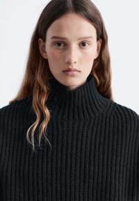 Black ribbed turtleneck sweater with a high collar, featuring a textured knit pattern and a relaxed fit. Soft, thick material.