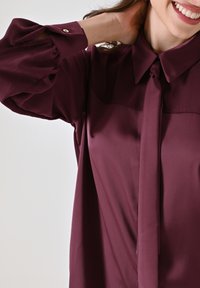 Blusa bordeaux con colletto a fiocco, maniche lunghe a sbuffo e dettagli con bottoni. Tessuto liscio con una leggera lucentezza e vestibilità su misura.