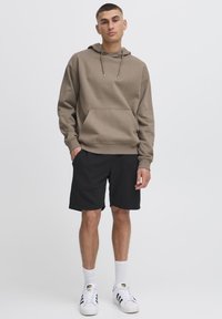 Hoodie marron avec une poche avant, assorti à un short noir, porté avec des chaussettes blanches et des baskets noires à rayures. Tissu lisse, coupe décontractée.