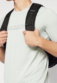 Czarny plecak z pikowanymi paskami, noszony na jasnoszarej dopasowanej koszuli z napisem "MAMMUT" w szarym kolorze. Ręce trzymające paski.