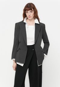 Mujer vistiendo una chaqueta de rayas finas gris oscuro sobre una camisa blanca y pantalones anchos negros, de pie frente a un fondo blanco plano.