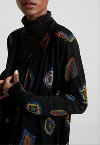 Schwarze Strickjacke mit bunten geometrischen Mustern, oversized Schnitt, langen Ärmeln und subtiler Textur mit kontrastierenden gerippten Bündchen.
