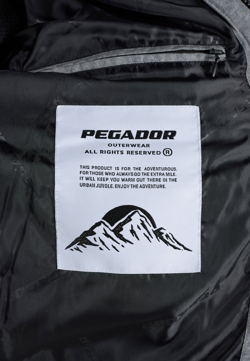 Etiqueta de la marca de ropa exterior White Pegador en tela negra con un gráfico de montaña y texto sobre calidez y aventura en una jungla urbana.