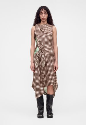 Femme aux cheveux bouclés portant une robe marron à pois sans manches avec un ourlet asymétrique et des bottes noires montant jusqu’aux genoux, debout sur un fond blanc.