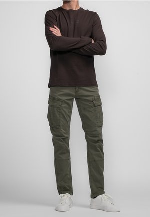 Mann mit verschränkten Armen, trägt ein dunkles Langarmshirt, olivgrüne Cargo-Hosen und weiße Turnschuhe vor einem schlichten grauen Hintergrund.