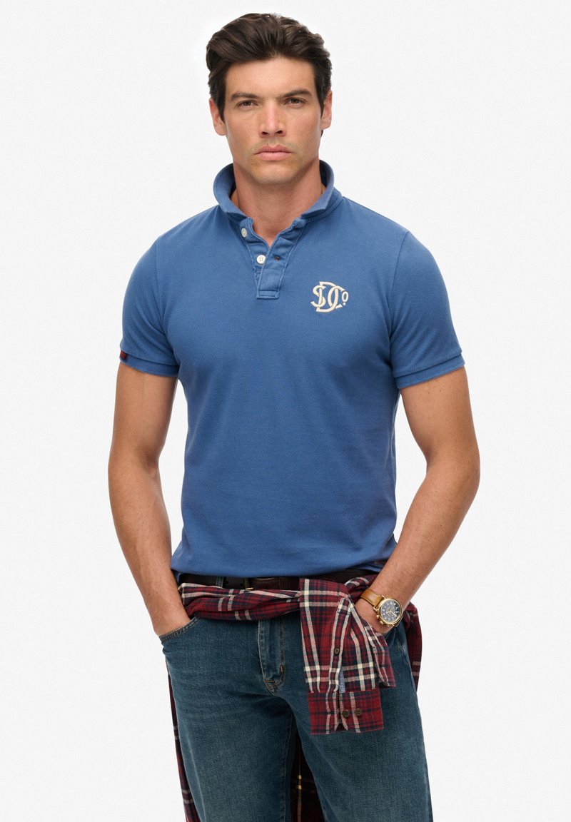Polo en coton bleu avec patte de boutonnage à deux boutons et logo brodé, associé à une flanelle à carreaux nouée à la taille et un jean en denim.