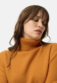 Oranžový rolák vyrobený z měkké úpletové tkaniny, s žebrovanou texturou na límci a manžetách. Hladký povrch s posunutými rameny.