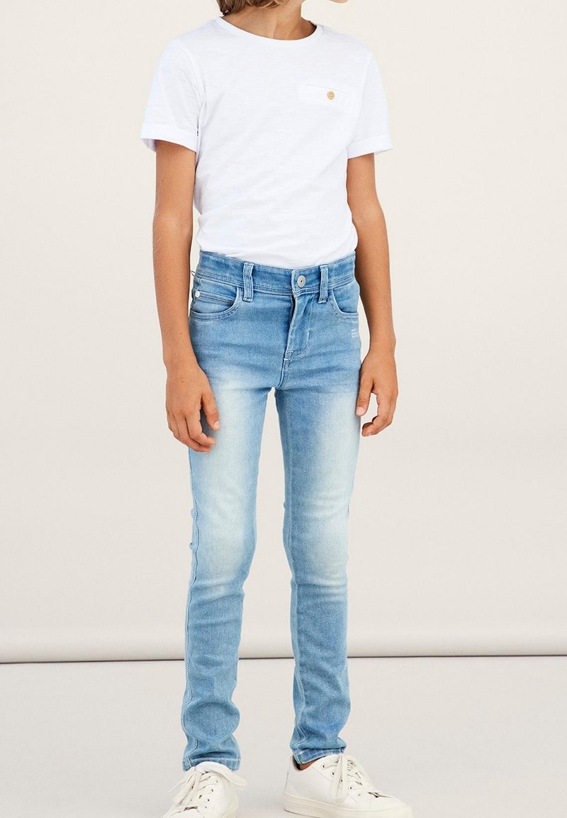 Enfant portant un t-shirt blanc à manches courtes, un jean skinny bleu clair délavé, et des baskets blanches sur un fond uni.
