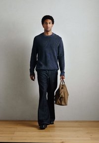 Camisola de malha azul escura com padrões canelados, jeans escuros evasés e uma mala tote castanha clara. O fundo é cinzento claro com chão de madeira.