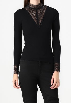 Haut noir cintré à manches longues avec des détails en dentelle sur le col montant, la poitrine et les poignets, porté avec un pantalon noir, visible des épaules jusqu'aux hanches.