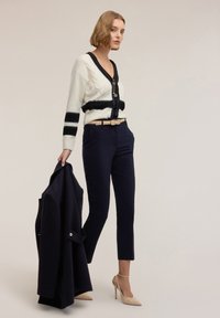 Biały, dziergany kardigan w czarne paski, zestawiony z granatowymi spodniami i beżowymi szpilkami. Modelka trzyma ciemny płaszcz. Prosty fason dekoltu.