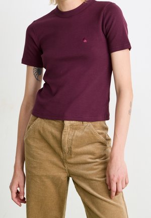 Persoon draagt een kortemouwen bordeaux geribd t-shirt en een lichtbruine hoge taille corduroy broek met hand in zak.